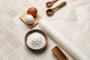 Baking paper with bowl of flour, eggs and spoons on beige tile background