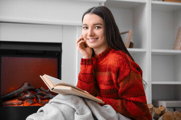Young woman in warm sweater reading book near fireplace at home