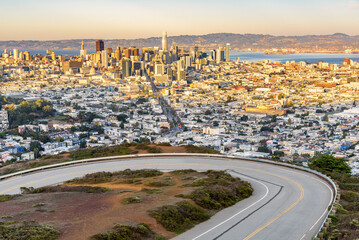 San Francisco skyline warmly lit by a setting sun in autumn. A winding road climbing Twin Peaks is in foreground. - Powered by Adobe