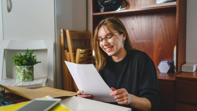 One young woman caucasian female student sitting at home holding envelope with mail letter happy exited smiling reading entry test results success and achievement concept real people copy space