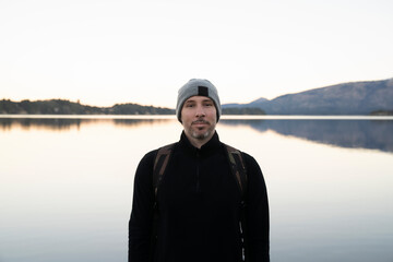 Portrait of a caucasian man wearing a hat, scarf and jacket with the lake and mountains in the background, at sunset. Beautiful landscape and sky reflection in the water surface.  - Powered by Adobe