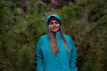 Hiking in the woods in a rainy day. Portrait of a young woman in her 20s, wearing a jacket, with the green forest canes in the background. 