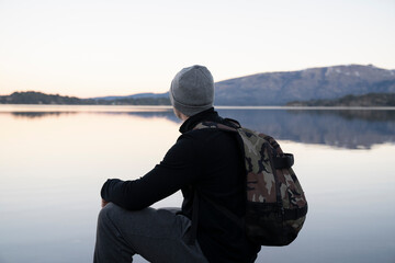 Tourism. Portrait of a young man wearing a hat and bag, contemplating the beautiful landscape, lake and mountains, at sunset. 