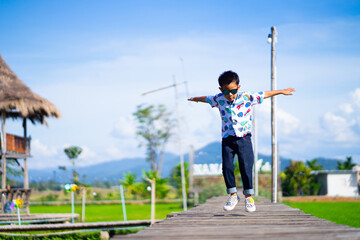 A boy jumping with his arms outstretched on a wooden bridge overlooking a rice field during a sunny afternoon