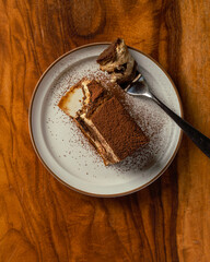 Tiramisu on round white plate with wooden background. Fork on a plate with a bite of tiramisu, lifestyle setup.