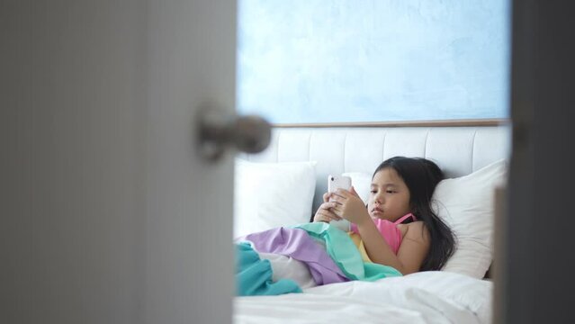 asian child addicted smartphone or kid girl sleep lying to pillow on bed and play watching mobile phone to hyperactive with parents secretly looking at daughter or baby behind ajar door in bedroom
