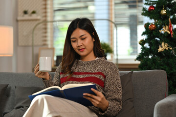 Millennial Asian woman in sweater reading a book and sipping a coffee on Christmas Day in the cozy...