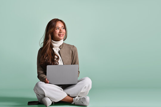Young Asian woman in winter outfit sit on the ground and use laptop on plain green background studio, Business and finance, Successful entrepreneurs, Sharing idea concept.