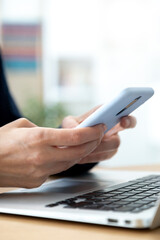 Vertical close-up of woman hands using mobile phone while working in the office with laptop. Selective focus on hands.