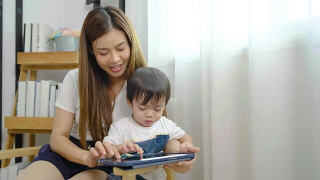 Young asian mother her little adorable son using tablet looking at computer screen enjoying watching funny social media video doing online shopping relaxing on sofa at home together
