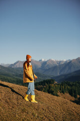 Fototapeta premium Woman autumn full-length walk on the hill smile with teeth and look at the mountains in a yellow raincoat and jeans happy hiking trip, freedom lifestyle 