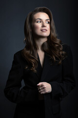 Fototapeta premium Fashion, business and make-up concept. Portrait of beautiful brunette woman with black suit and big wavy hair in dark studio background. Model looking aside the camera. Toned image with blue color