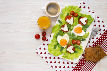 Healthy festive breakfast on Valentine's Day. plate of heart-shaped fried eggs, tomatoes, cheese, coffee and juice on a white wooden table covered with a napkin. View from above. Space for text.