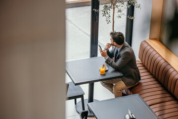 Pensive businessman in a gray suit, checking his emails on his mobile phone, drinking a coffee and fresh orange juice in the local cafe.