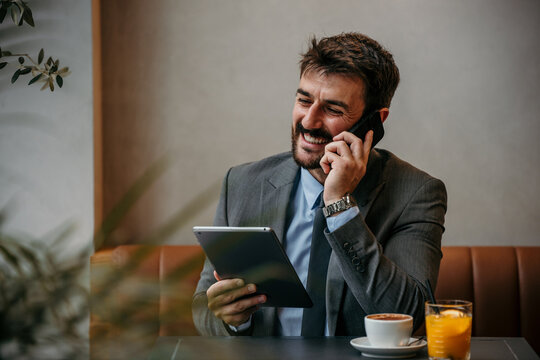 Smiling executive sitting in the lounge bar, talking on the mobile phone and holding a digital tablet.