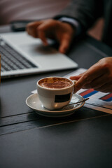 A businessman working in the office has a laptop, papers, and a coffee on his desk, focus on one hand taking a coffee, while the other hand is typing on his laptop.