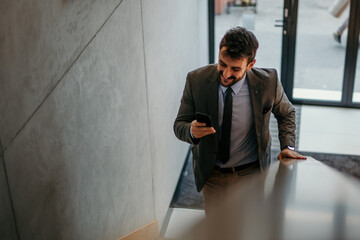 A cheerful executive climbs up the stairs in the office building, looking at his phone and texting.