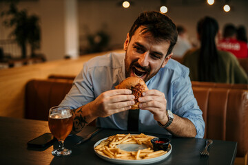 Man in a business shirt and tie with rolled up sleeves sitting in the pub, holding and enjoying a burger and beer.