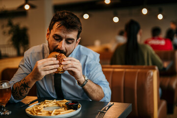 Man in a business shirt and tie with rolled up sleeves sitting in the pub, holding and enjoying a burger and beer.