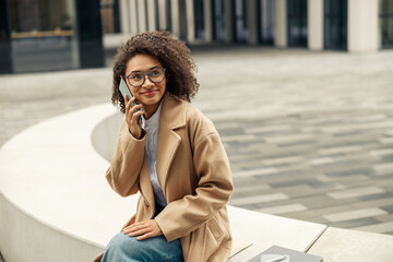 Smiling afro american businesswoman talking phone while sitting on background of modern building 