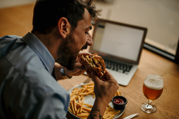 Close up image of a handsome bearded businessman eating his burger, drinking a beer, and working remotely on a laptop.