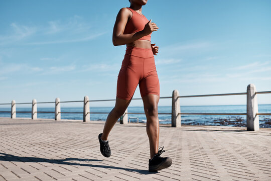Black woman, legs or running by city beach, ocean or sea in healthcare workout, cardiovascular wellness training or marathon exercise. Runner, sports athlete or personal trainer on promenade fitness