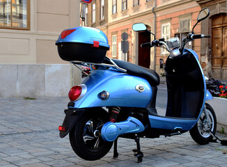 electric scooter. small light blue moped with storage box. old  urban historic city setting. light gray cobblestone pavement. travel and tourism to europe concept - obrazy, fototapety, plakaty