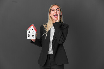 Blonde Uruguayan girl holding a house toy isolated on black background shouting with mouth wide open