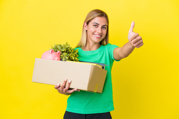 Young Uruguayan girl making a move while picking up a box full of things isolated on yellow background with thumbs up because something good has happened