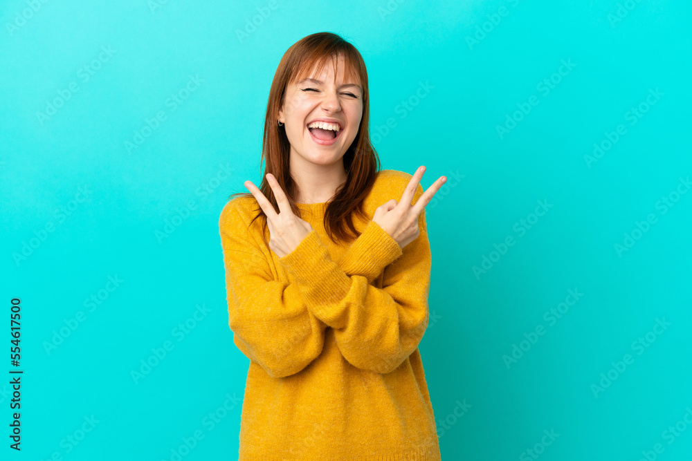 Wall mural Redhead girl isolated on blue background smiling and showing victory sign