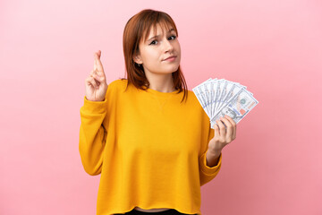 Redhead girl taking a lot of money isolated on pink background with fingers crossing and wishing the best