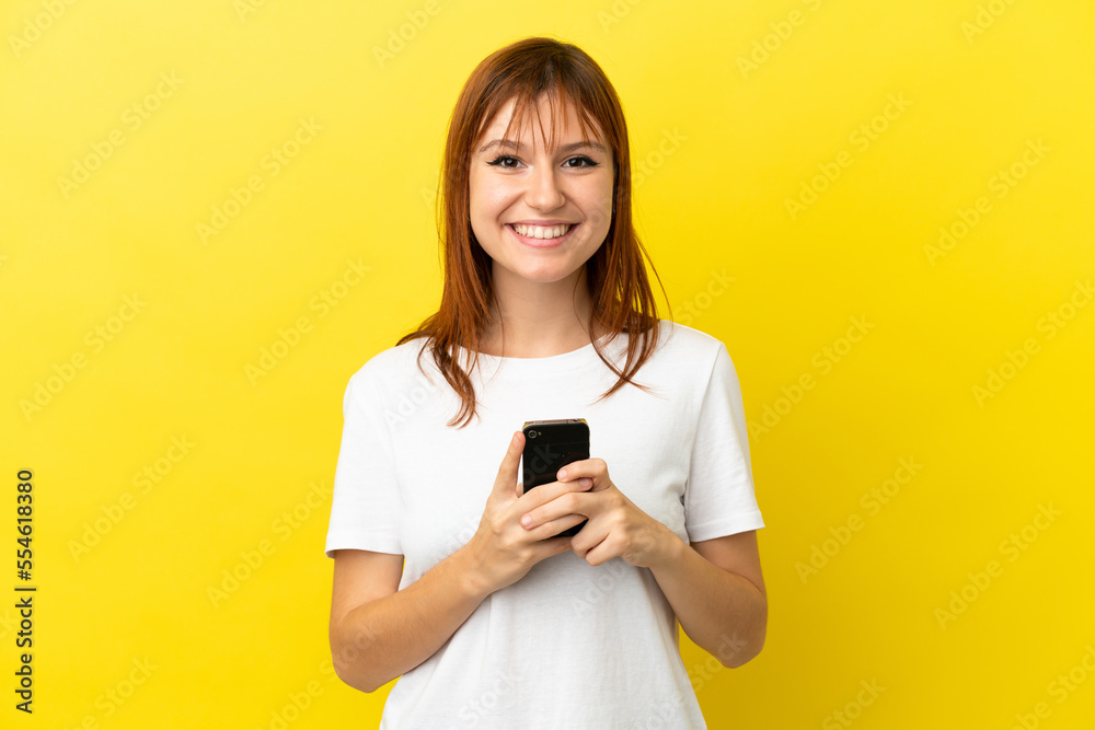 Wall mural Redhead girl isolated on yellow background looking at the camera and smiling while using the mobile