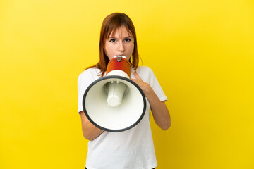 Redhead girl isolated on yellow background shouting through a megaphone with surprised expression