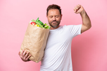 Middle age man holding a grocery shopping bag isolated on pink background doing strong gesture