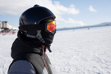 Fototapeta na wymiar Girl or boy in ski helmet, sunscreen mask and balaclava close up stands against the backdrop of snow-covered mountain ski slope and a cloudy sky. Winter. Sport and travel content 