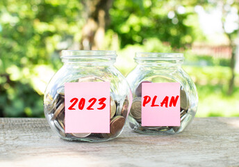 Glass jars with coins and the inscriptions 2023 and PLAN stand on a wooden table. Investment budget.