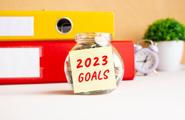 A glass jar with coins is on the work table. There is a sticker on the bank with the text 2023 GOALS.