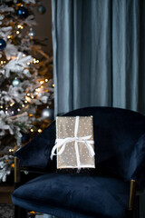 A gift in a silver wrapper with a white ribbon on a velvet blue chair against the background of a Christmas tree..