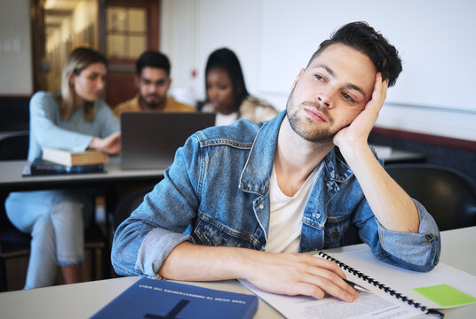 Education, university and bored student in classroom thinking studying for exam. Sad young man at college, boredom and stress, depression or anxiety with book and academic students at desk on campus.