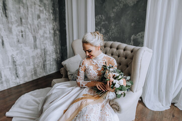 Fototapeta na wymiar A beautiful young bride model blonde in a long lace dress sits on a sofa in the interior with a bouquet in her hands. Wedding photography, portrait.