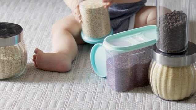 baby playing with jars with grains spices ingredients for kitchen home food preparing.plastic or glass jars on floor carpet unrecognizable toddler legs infant.millet, linseed, cuscus peppercorns