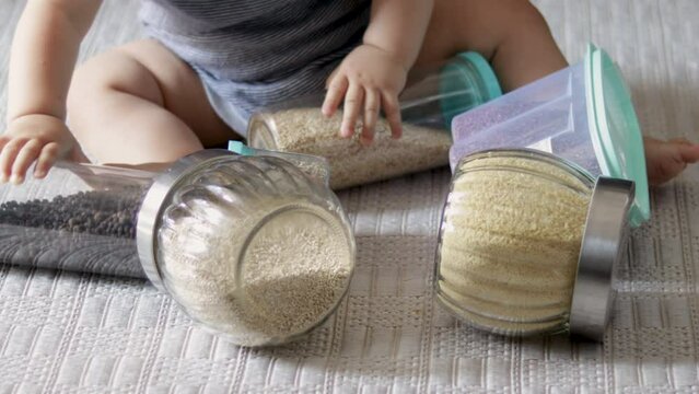 baby playing with jars with grains spices ingredients for kitchen home food preparing.plastic or glass jars on floor carpet unrecognizable toddler legs infant.millet, linseed, cuscus peppercorns