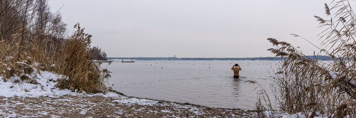 Ein junger Mann geht mit im Winter Badehose und Mütze in einem See schwimmen, Panorama