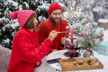 Fototapeta na wymiar Man and woman in red winter clothes have fun while eating fondue, sitting together by the table outdoors at snowy garden. Young family celebrating winter holidays