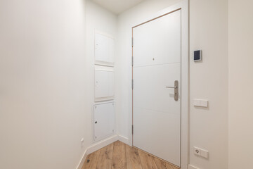 White closed wooden front door inside the apartment leading to the street. Apartment with white walls and parquet floors. The door has a built-in metal handle with a lock for opening and closing.