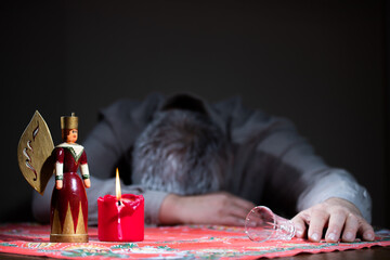A lonely, unhappy man sits sadly at a table. On the table are a Christmas angel, a candle and a shot glass.
