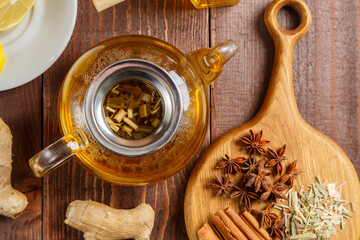 Transparent teapot of lemongrass tea next to anise, ginger and cinnamon on a round wooden board.