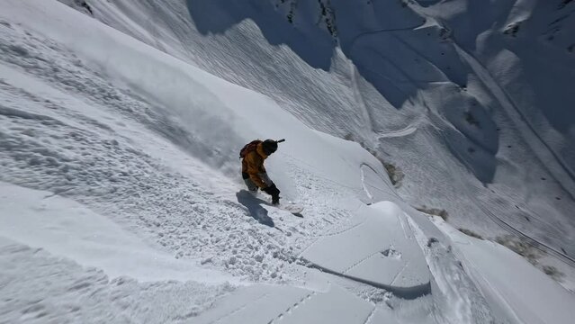 Male snowboarder freeride riding from mountain summit drop snow splash extreme sport tourism aerial view. FPV drone shot active man snowboard rider enjoy adrenaline freedom winter cliff landscape 4k