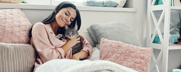 Attractive young woman in pajamas petting a cat and smiling while resting in bed at home