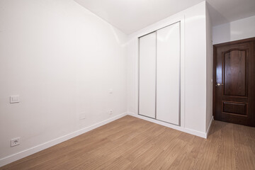 A room with a custom built-in wardrobe with white sliding doors, white painted smooth walls and dark wooden doors